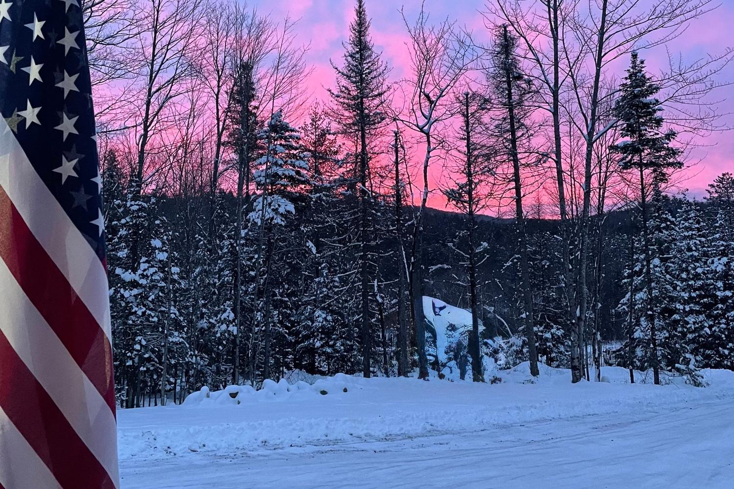 An American flag in the foreground and a pink winter sunset behind snow-covered spruce and bare trees above a snowy road in northern Vermont